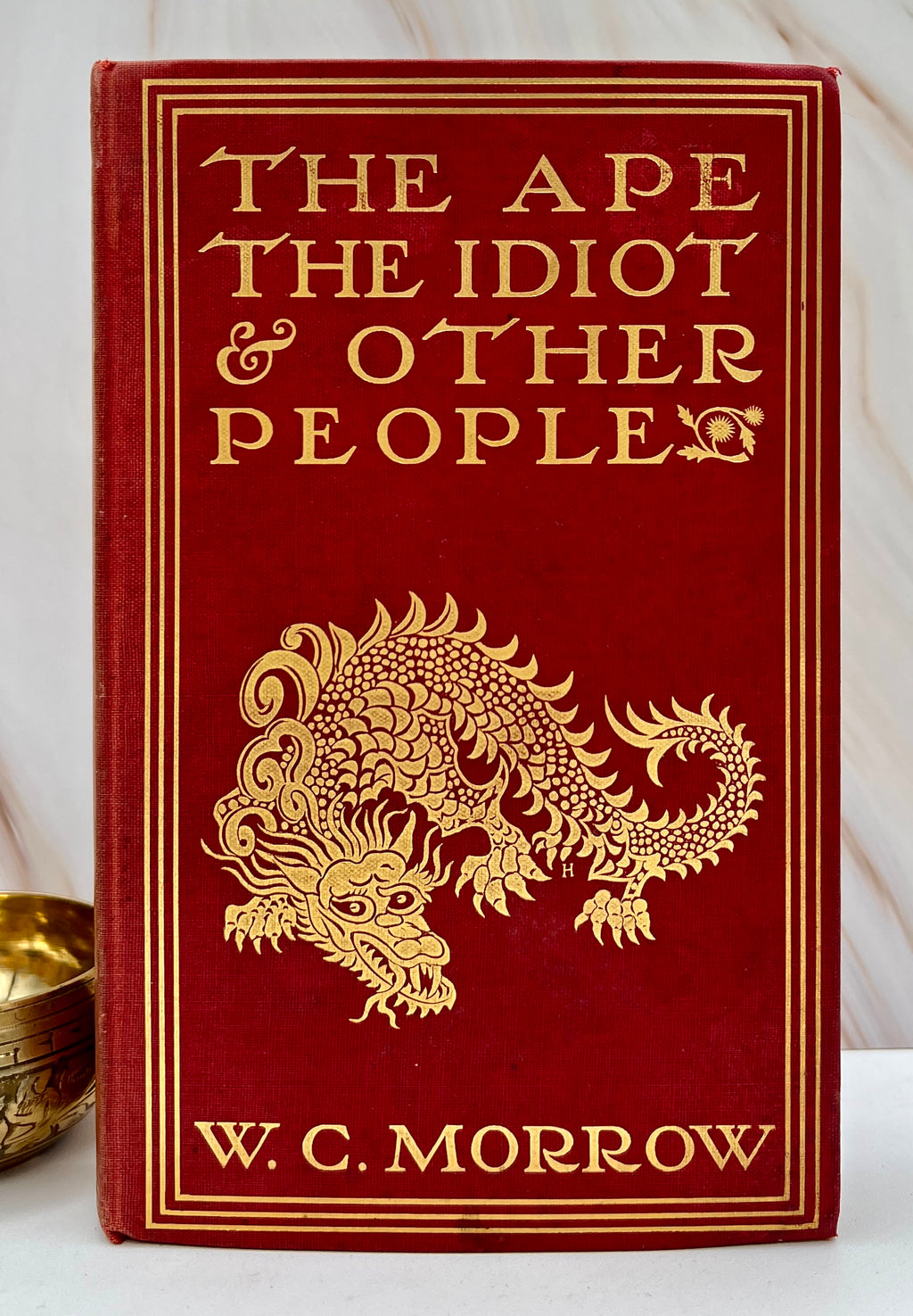 Antique red cloth hardcover book titled The Ape, the Idiot & Other People by W. C. Morrow, featuring an ornate gilt-stamped mythical dragon and gold lettering. - Photo 1