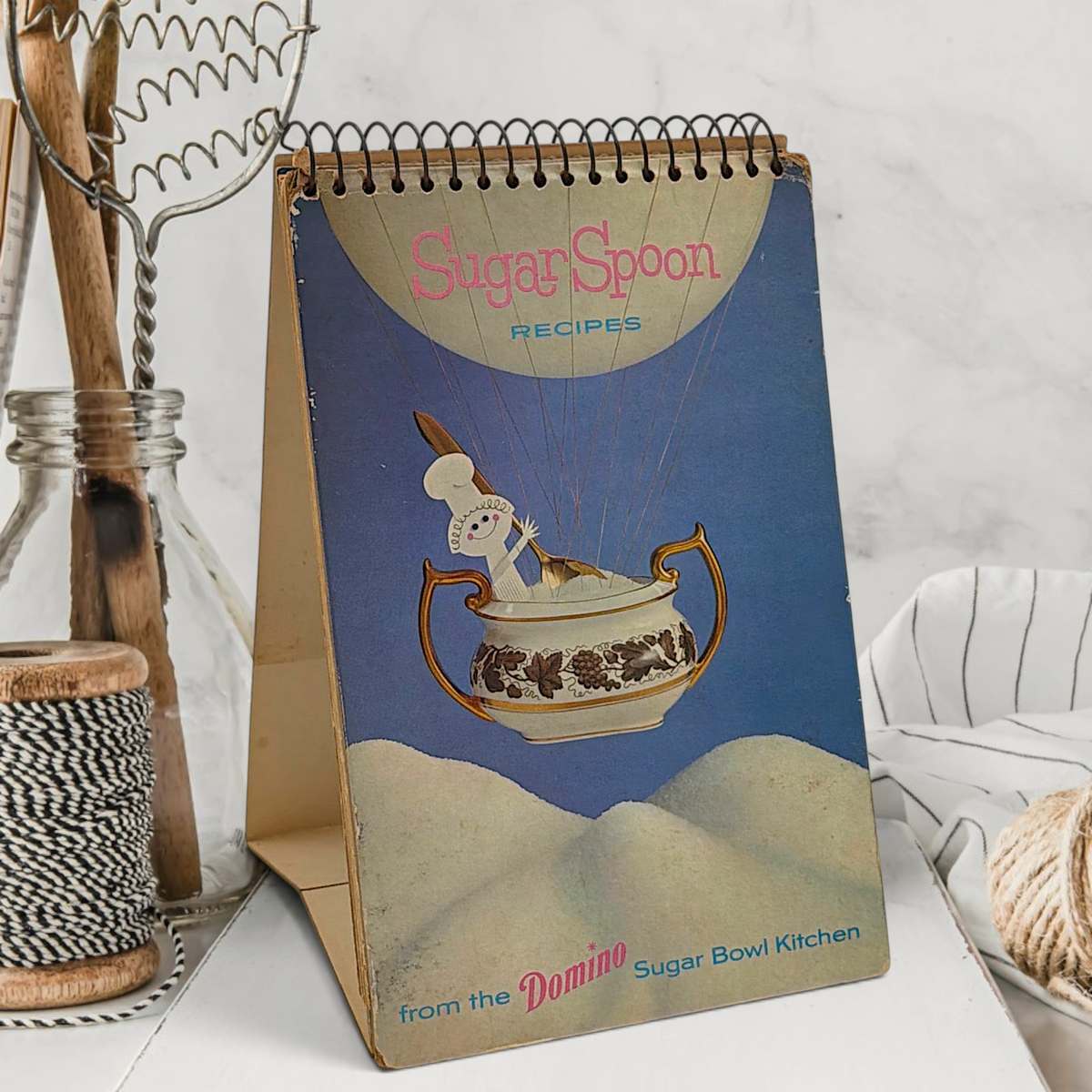 A vintage spiral-bound recipe easel stand featuring an illustration of a tiny chef piloting an antique floral sugar bowl hot air balloon over sugar dunes under a blue sky. - Photo 1