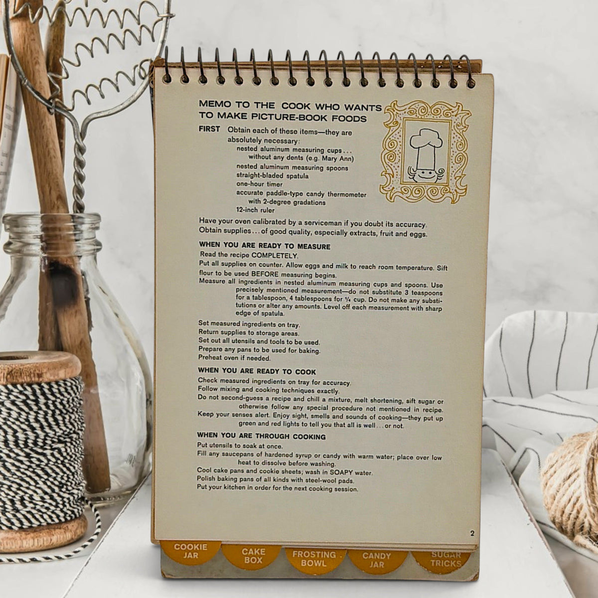 A vintage spiral-bound recipe easel stand featuring an illustration of a tiny chef piloting an antique floral sugar bowl hot air balloon over sugar dunes under a blue sky. - Photo 3