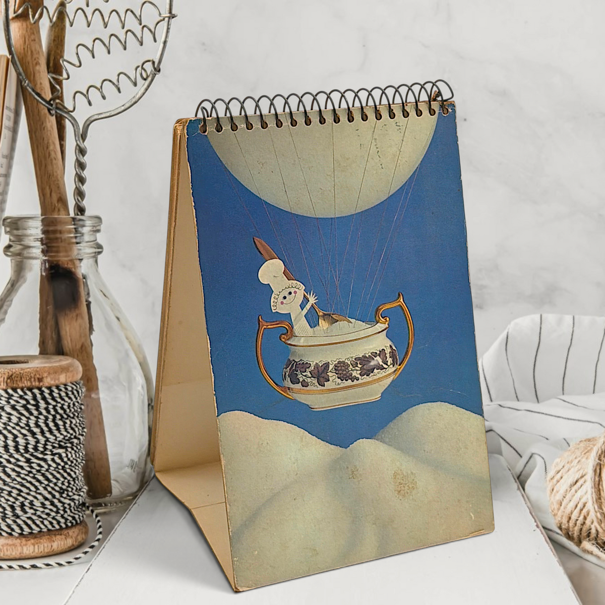 A vintage spiral-bound recipe easel stand featuring an illustration of a tiny chef piloting an antique floral sugar bowl hot air balloon over sugar dunes under a blue sky. - Photo 6