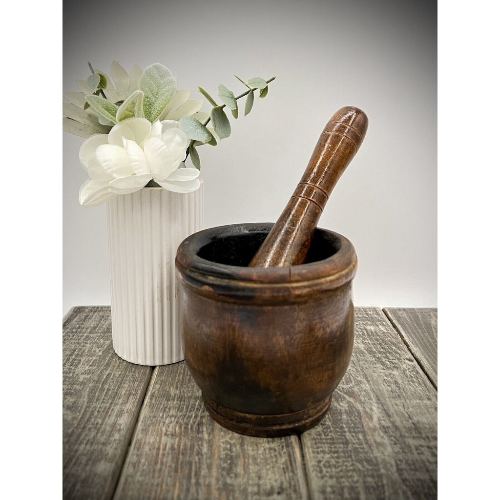 Antique wooden mortar and pestle with a tall worn pestle, set on a weathered table beside a white floral vase.