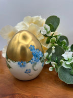 Decorative egg with floral design and gold finish on a wooden surface with flowers.