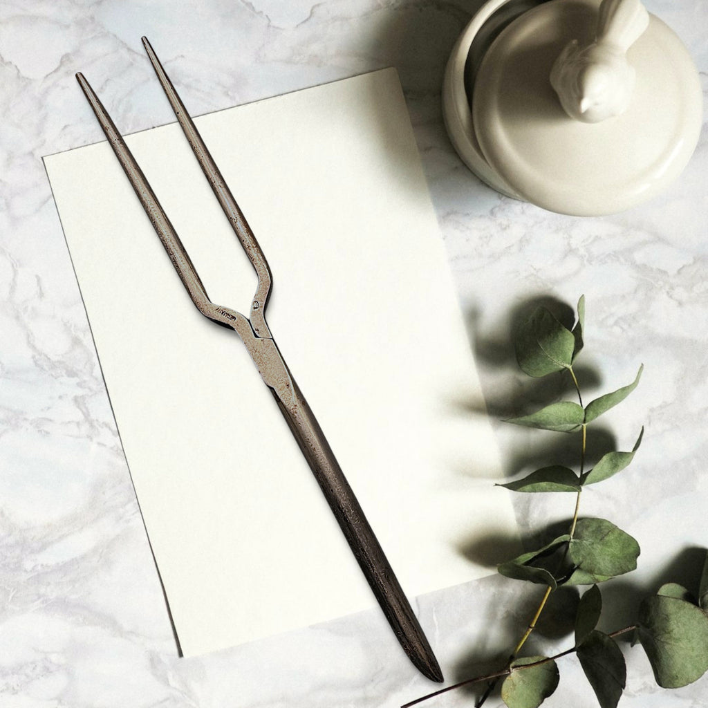 Two antique forged steel Marcel curling iron tongs with tapered points resting diagonally on white paper beside a dried eucalyptus branch. - Photo 1