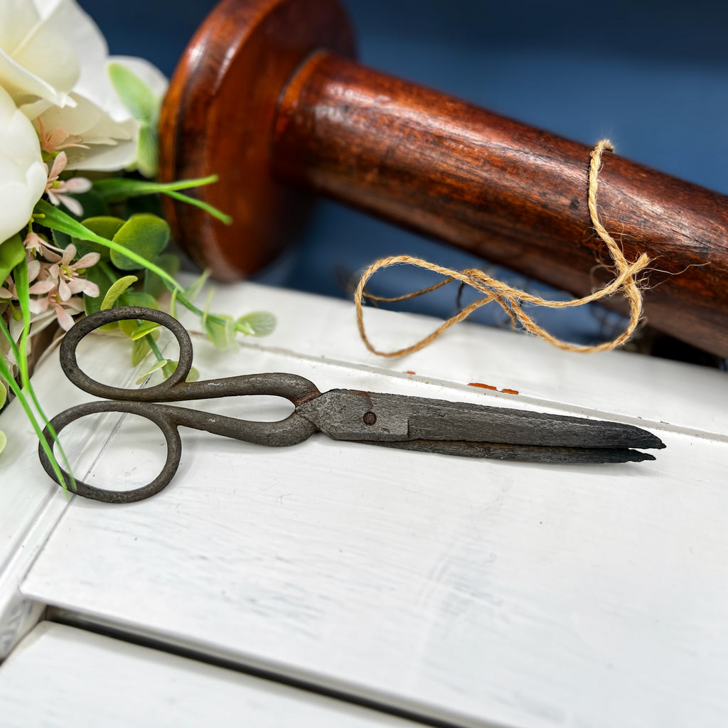 Antique hand-forged iron shears with rounded finger loops and tapered blades resting on a white surface beside a wooden textile bobbin. - Photo 1