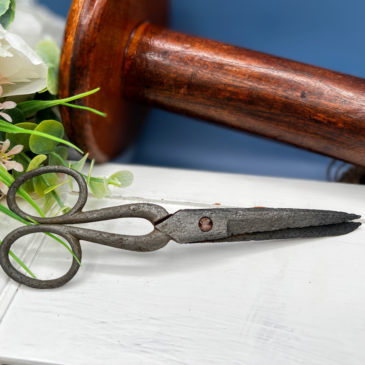 Antique hand-forged iron shears with rounded finger loops and tapered blades resting on a white surface beside a wooden textile bobbin. - Photo 6