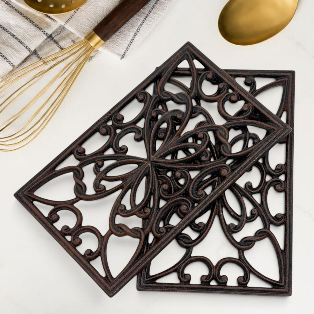 A rectangular black cast iron trivet displaying an ornate openwork scrollwork pattern with symmetrical botanical motifs, resting on a white marble countertop. - Photo 1