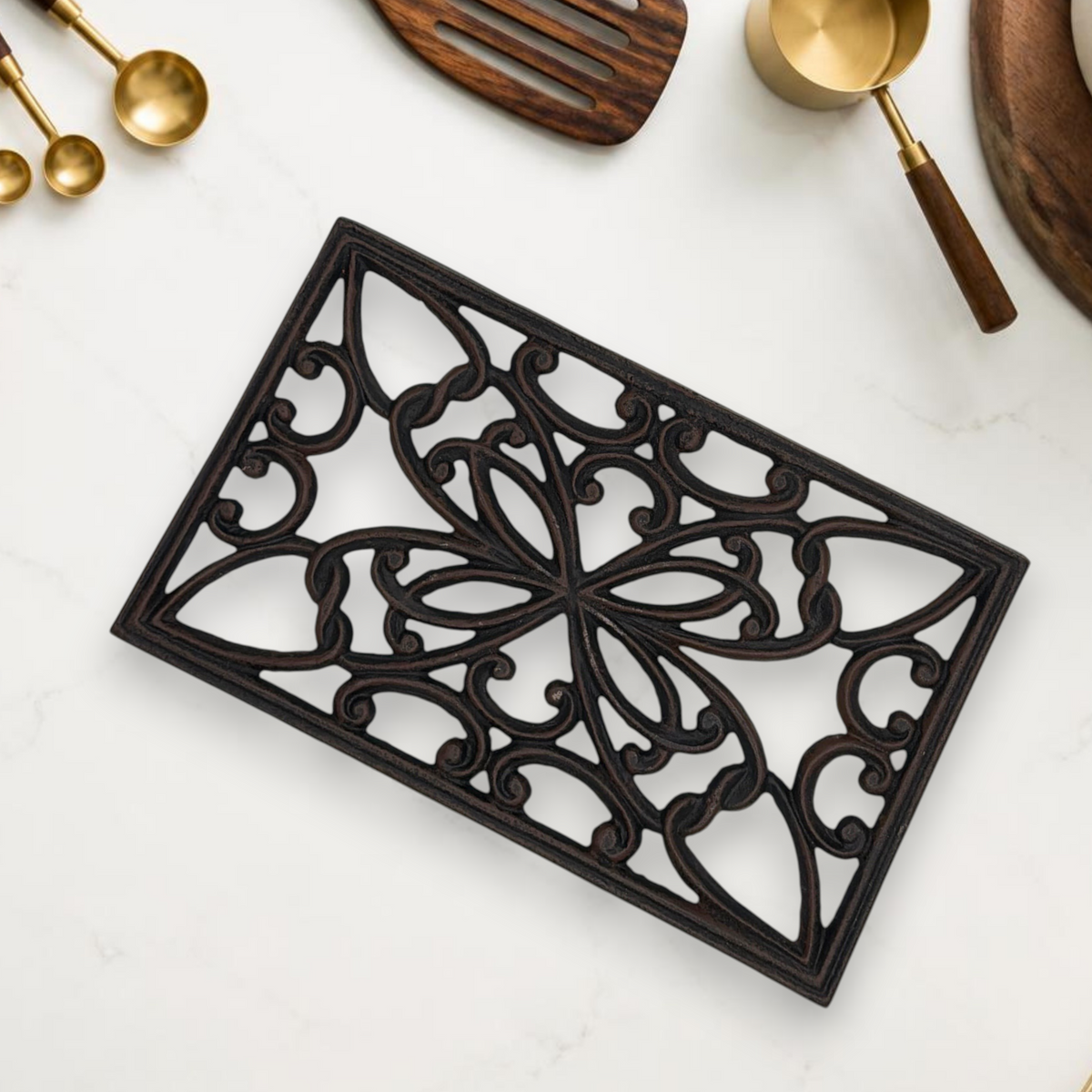 A rectangular black cast iron trivet displaying an ornate openwork scrollwork pattern with symmetrical botanical motifs, resting on a white marble countertop. - Photo 2
