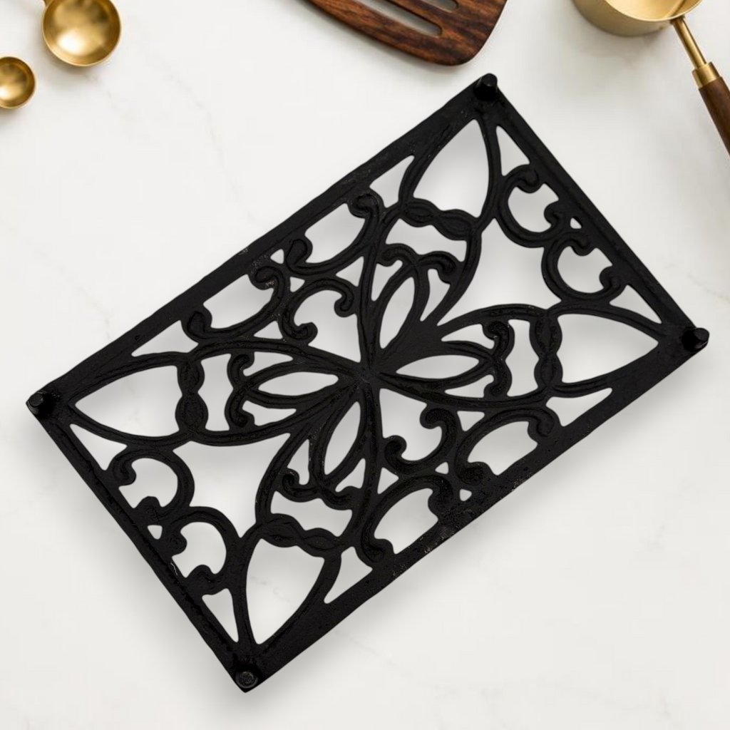 A rectangular black cast iron trivet displaying an ornate openwork scrollwork pattern with symmetrical botanical motifs, resting on a white marble countertop. - Photo 3
