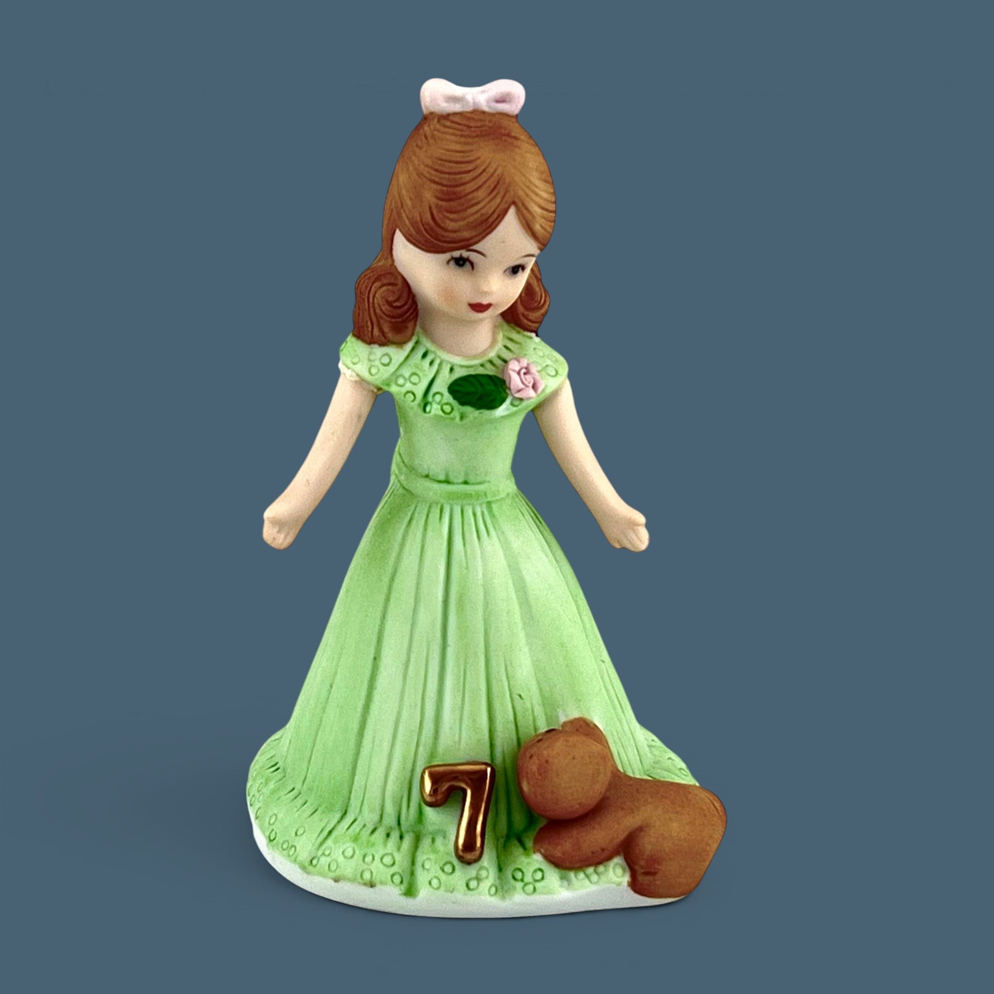 An Enesco Growing Up Birthday Girls porcelain figurine for age seven, featuring a young girl with light brown hair in a pastel green gown with a gold number seven and a brown teddy bear by her side. - Photo 1