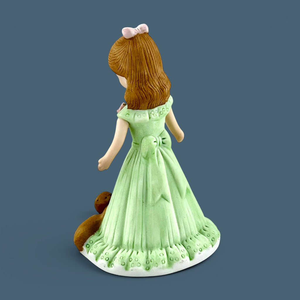 An Enesco Growing Up Birthday Girls porcelain figurine for age seven, featuring a young girl with light brown hair in a pastel green gown with a gold number seven and a brown teddy bear by her side. - Photo 2
