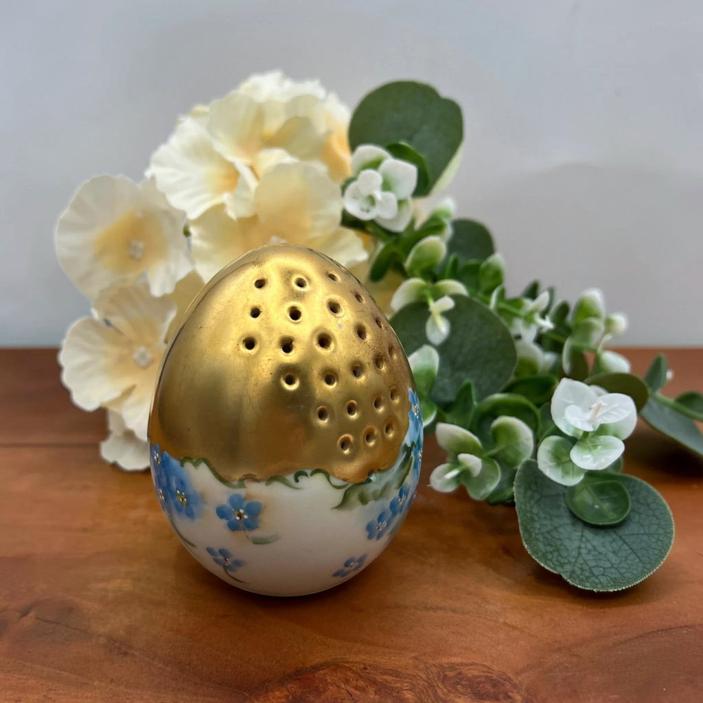 An antique porcelain egg-shaped shaker featuring hand-painted blue forget-me-not flowers along the base and heavy, lustrous gold gilding across the top, set against a background of cream flowers and green foliage. - Photo 1