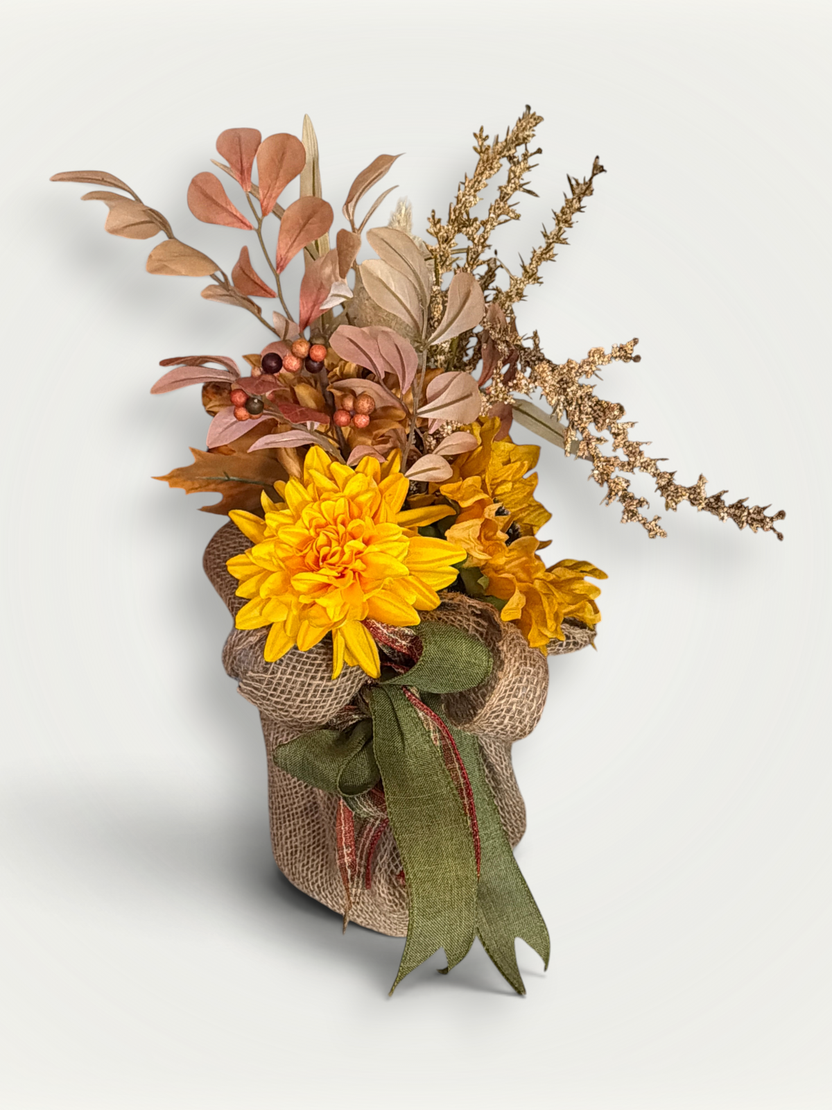 Artificial autumn floral arrangement featuring a bright yellow dahlia, peach-toned leaves, goldenrod sprigs, and dark berries set in a burlap-wrapped pot tied with a green ribbon.
