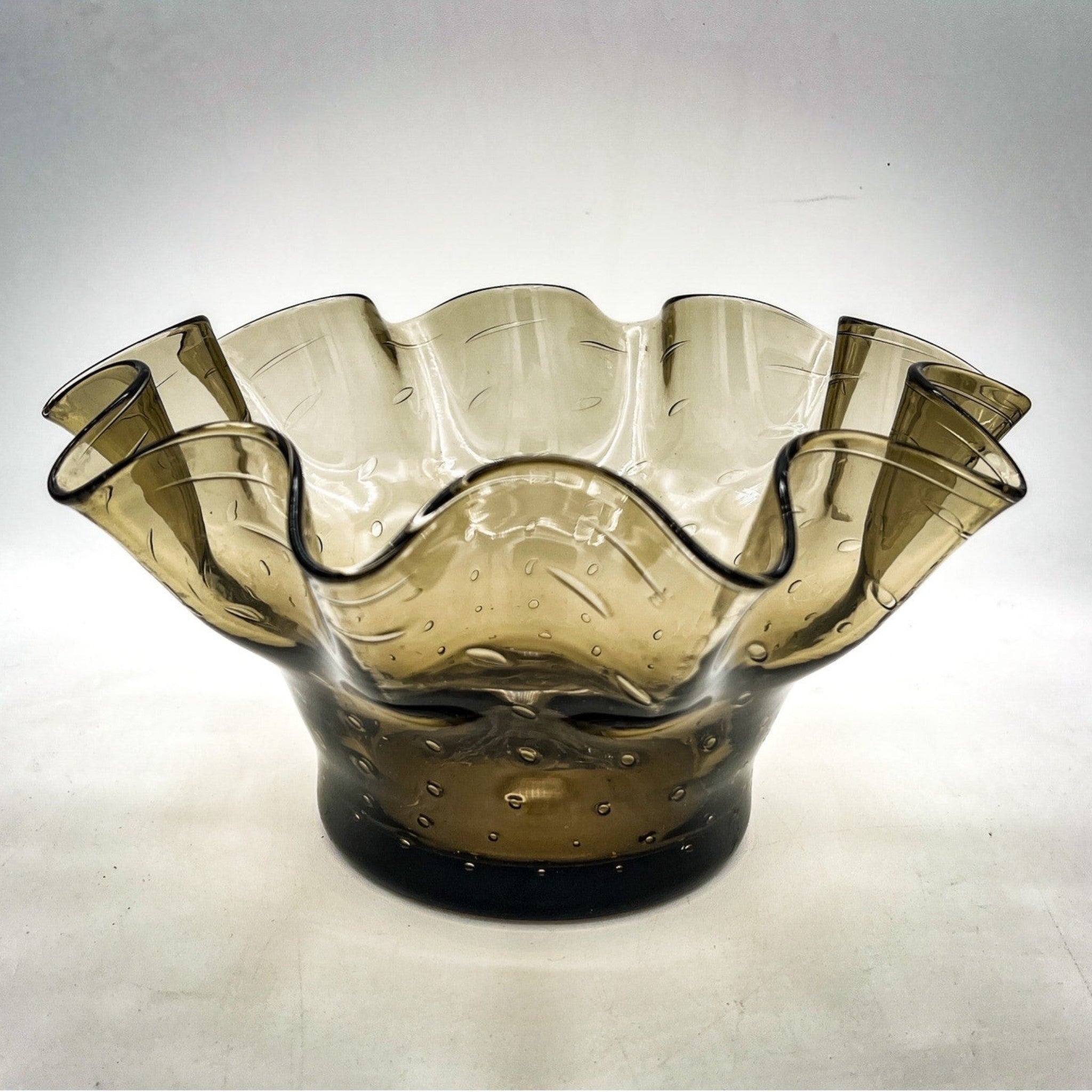 A smoky gray hand-blown art glass console bowl with a thick base featuring a neat row of controlled bubbles and a wide, undulating crimped ruffled rim flared outward against a white background. - Photo 1