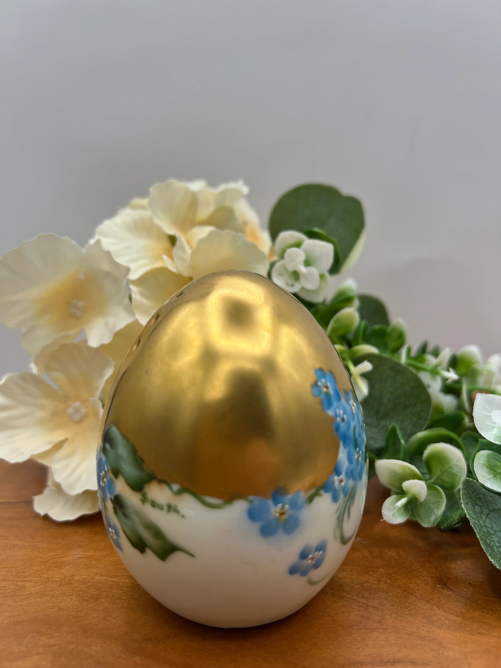 Decorative egg with gold top and floral design on a wooden surface with flowers in the background