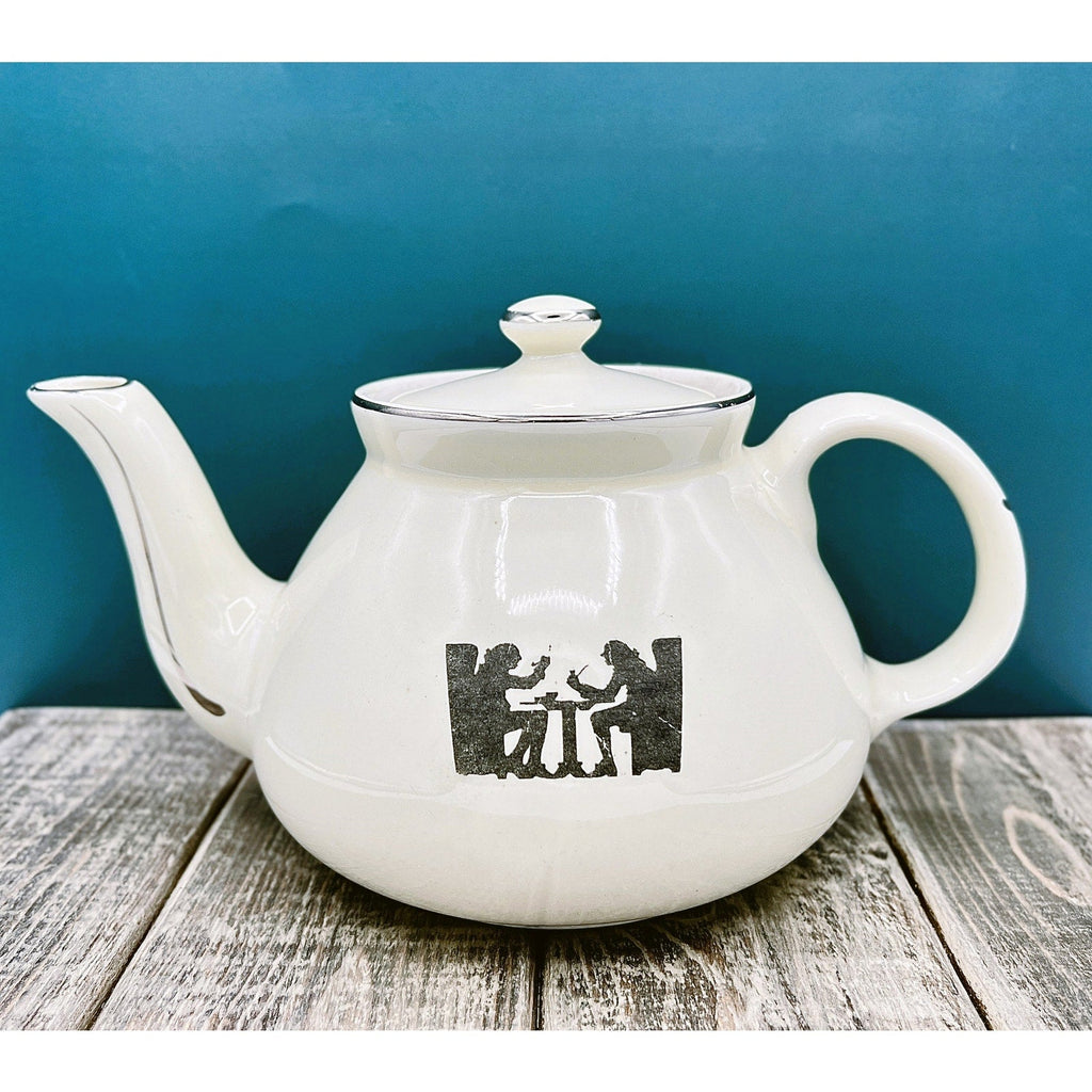 A warm cream ceramic teapot with a black silhouette tavern scene decal, showcasing a classic Art Deco streamlined design and platinum luster accents on the sweeping handle and spout. - Photo 1