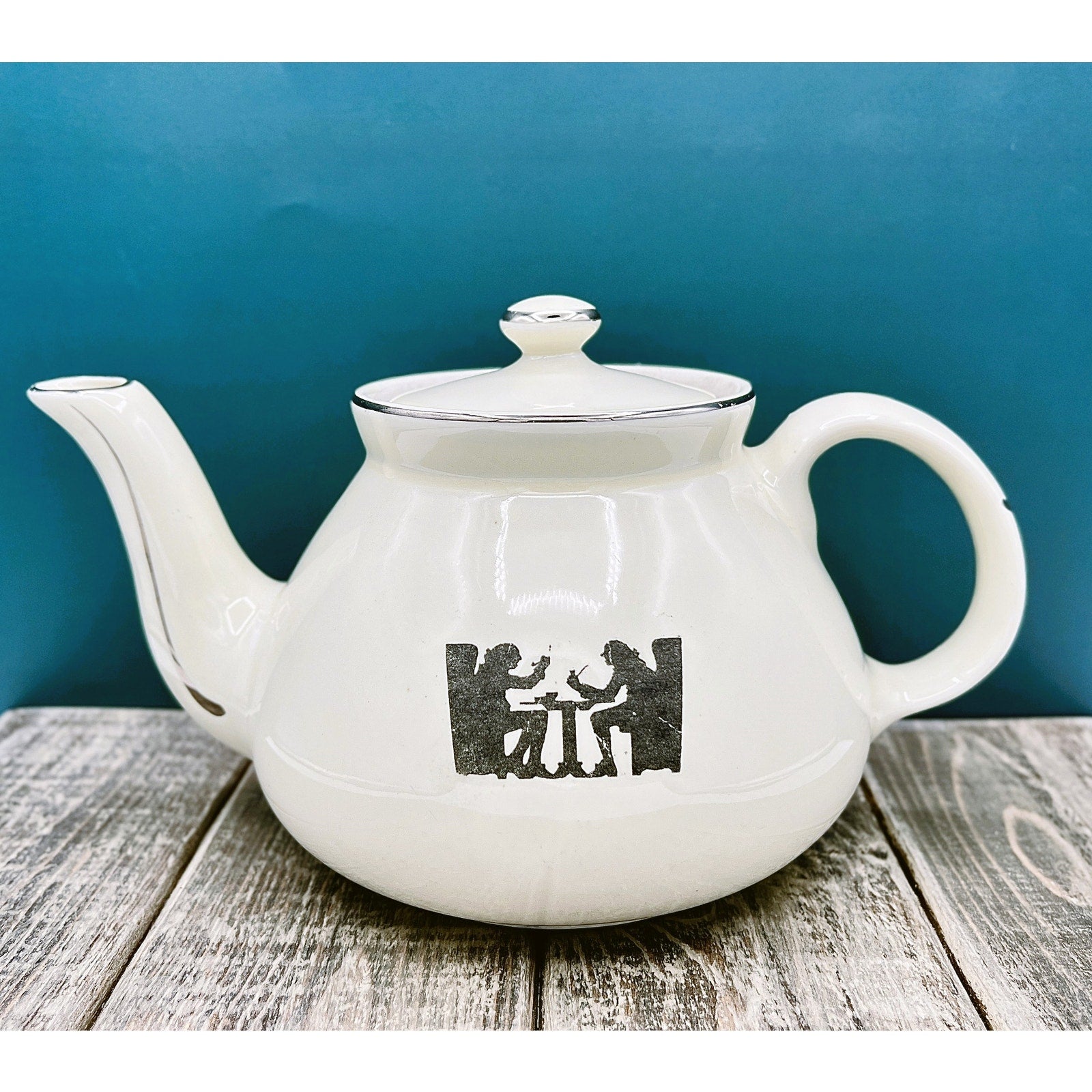 A warm cream ceramic teapot with a black silhouette tavern scene decal, showcasing a classic Art Deco streamlined design and platinum luster accents on the sweeping handle and spout. - Photo 1