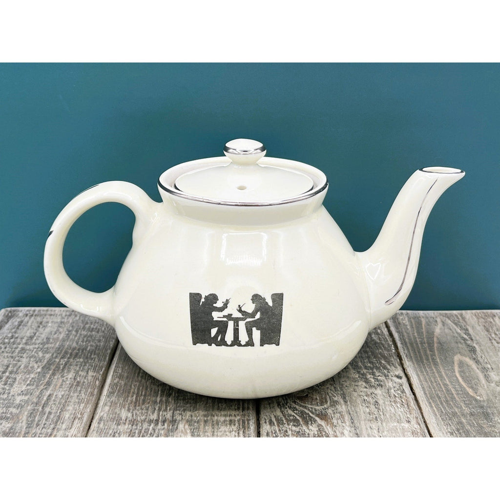A warm cream ceramic teapot with a black silhouette tavern scene decal, showcasing a classic Art Deco streamlined design and platinum luster accents on the sweeping handle and spout. - Photo 2