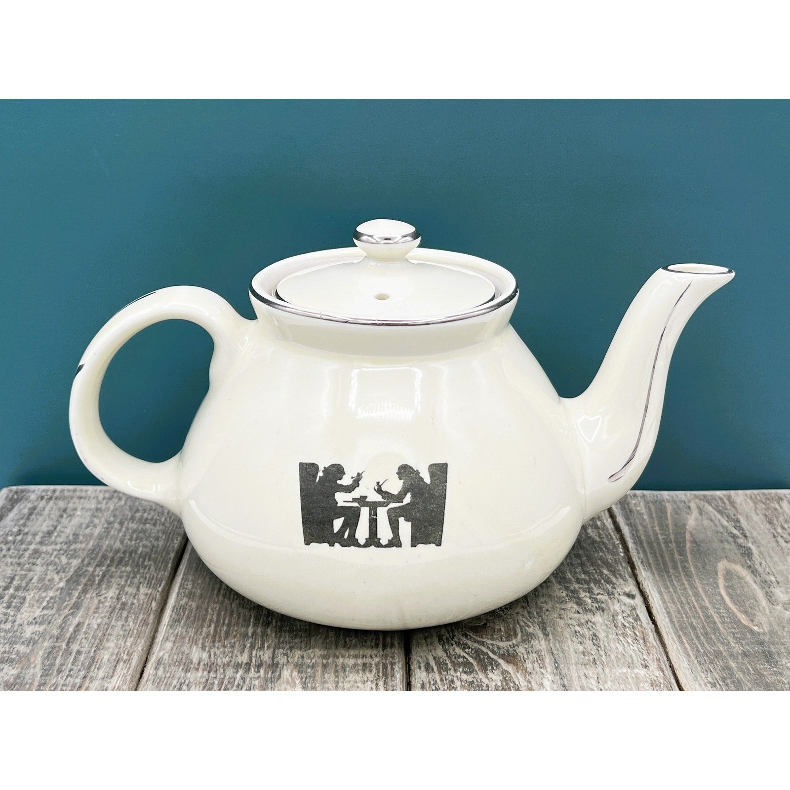A warm cream ceramic teapot with a black silhouette tavern scene decal, showcasing a classic Art Deco streamlined design and platinum luster accents on the sweeping handle and spout. - Photo 2