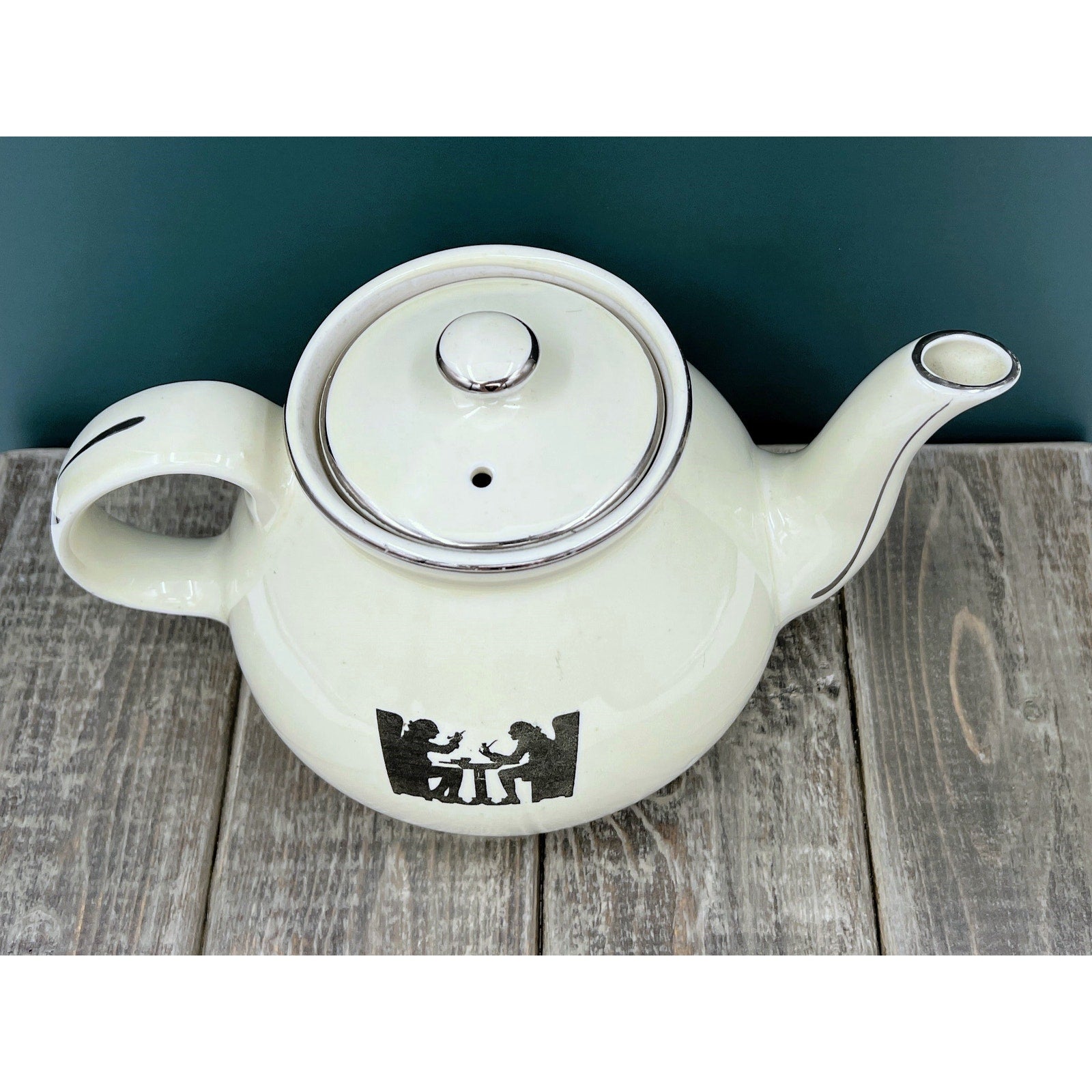 A warm cream ceramic teapot with a black silhouette tavern scene decal, showcasing a classic Art Deco streamlined design and platinum luster accents on the sweeping handle and spout. - Photo 3