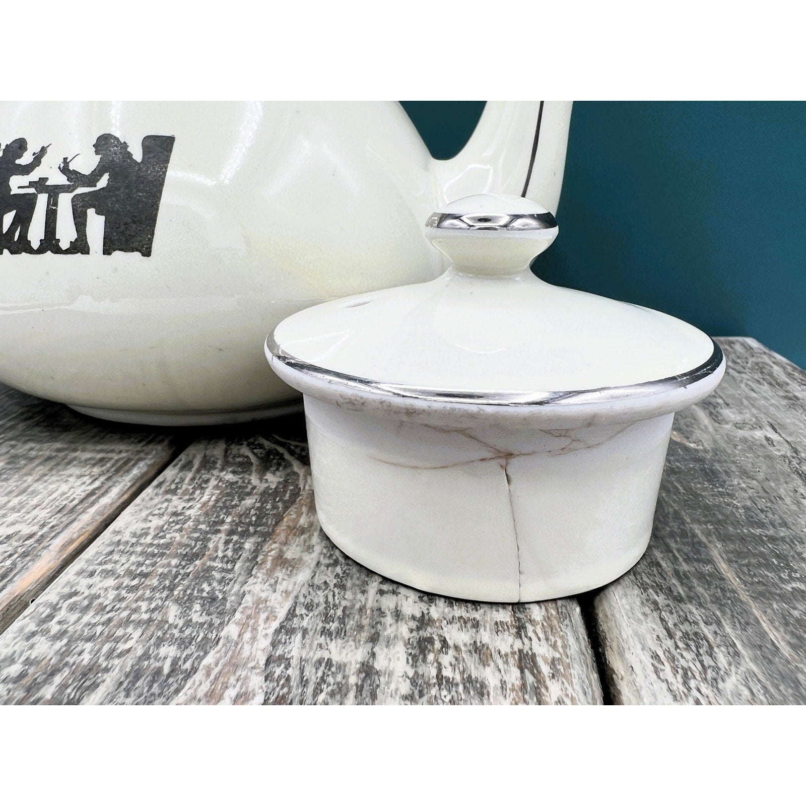 A warm cream ceramic teapot with a black silhouette tavern scene decal, showcasing a classic Art Deco streamlined design and platinum luster accents on the sweeping handle and spout. - Photo 7