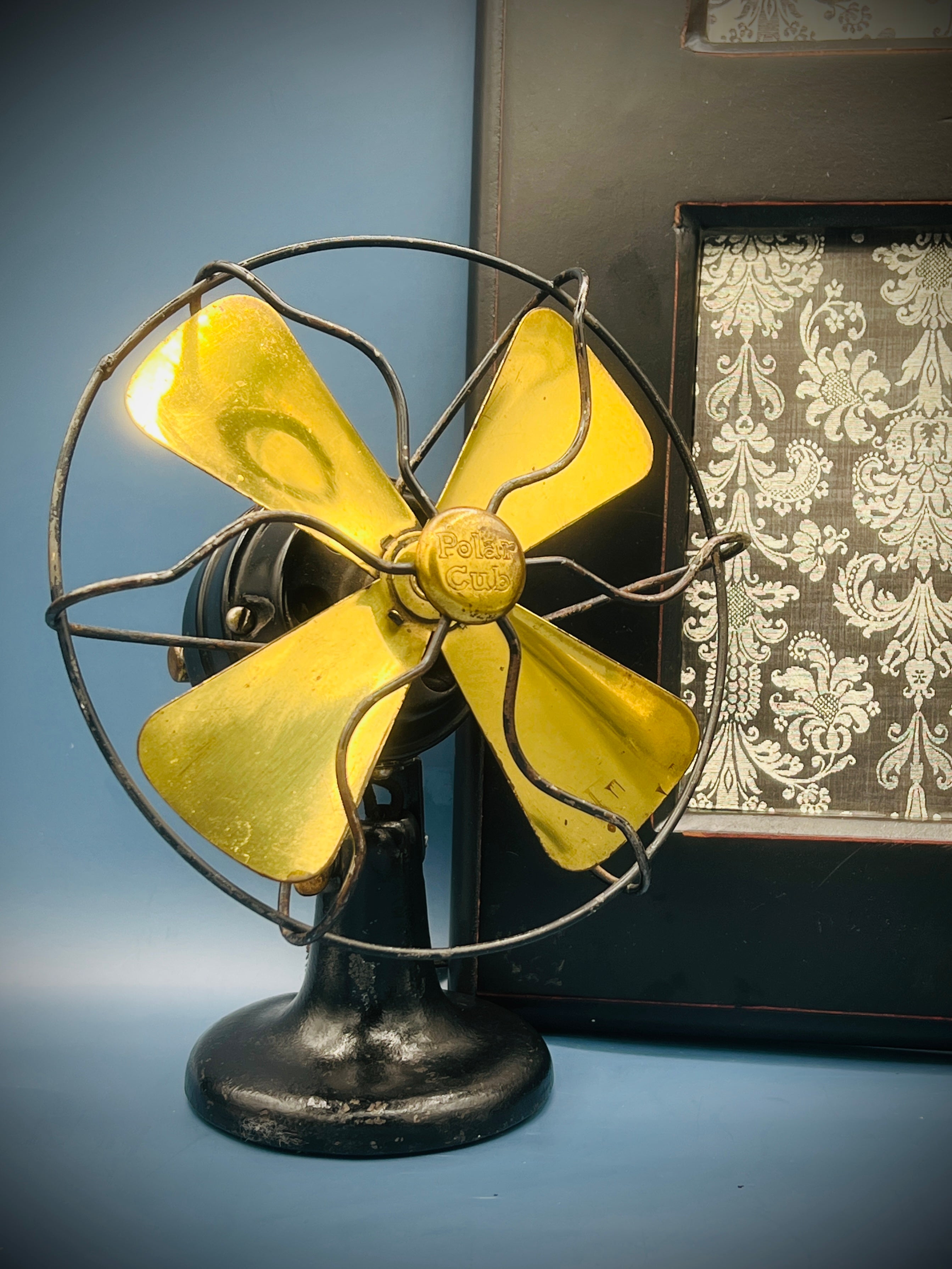 A vintage A.C. Gilbert Polar Cub electric fan featuring four brass blades, a wire cage guard, and a black painted metal base set against a patterned background. - Photo 1