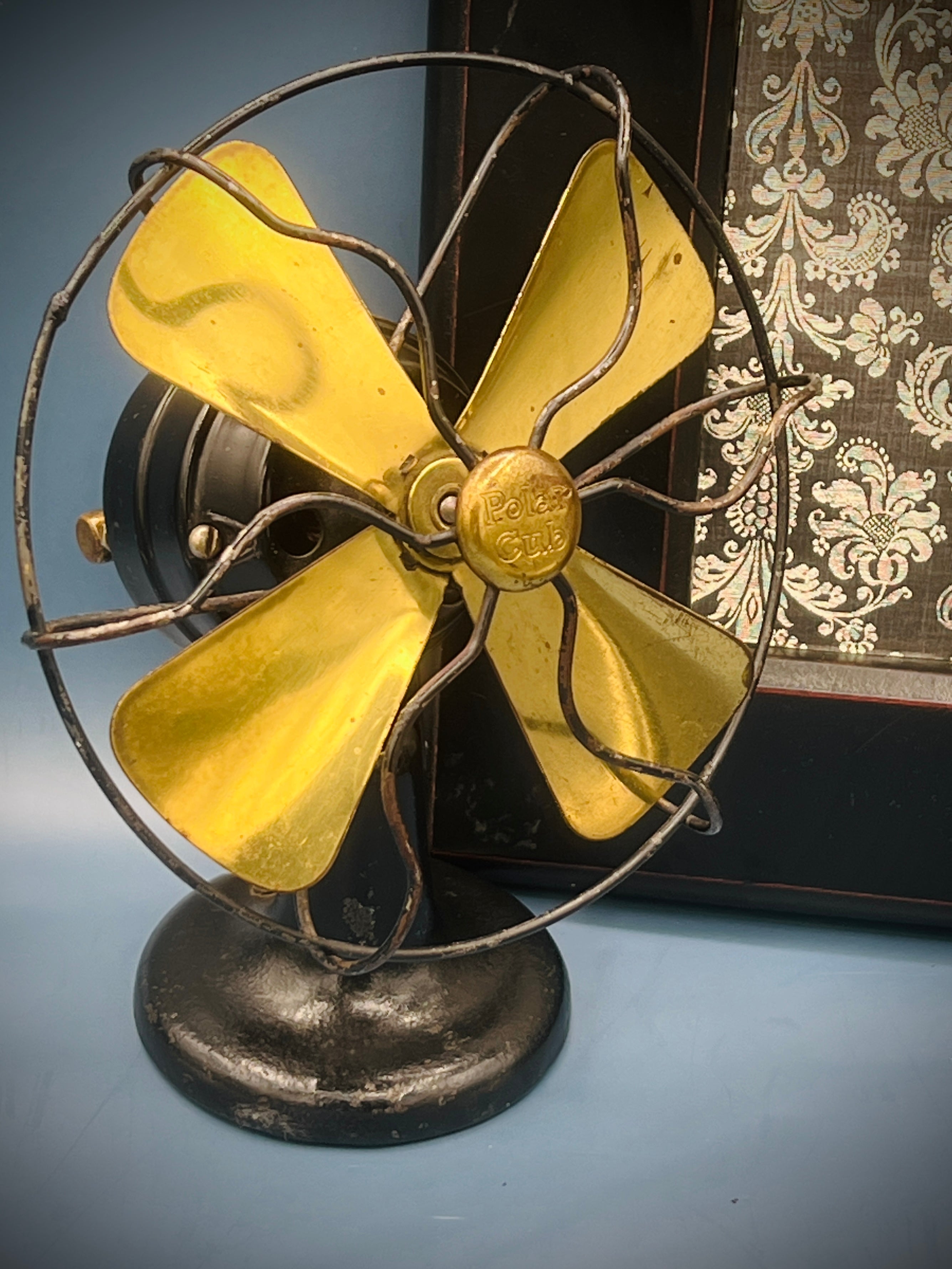 A vintage A.C. Gilbert Polar Cub electric fan featuring four brass blades, a wire cage guard, and a black painted metal base set against a patterned background. - Photo 2