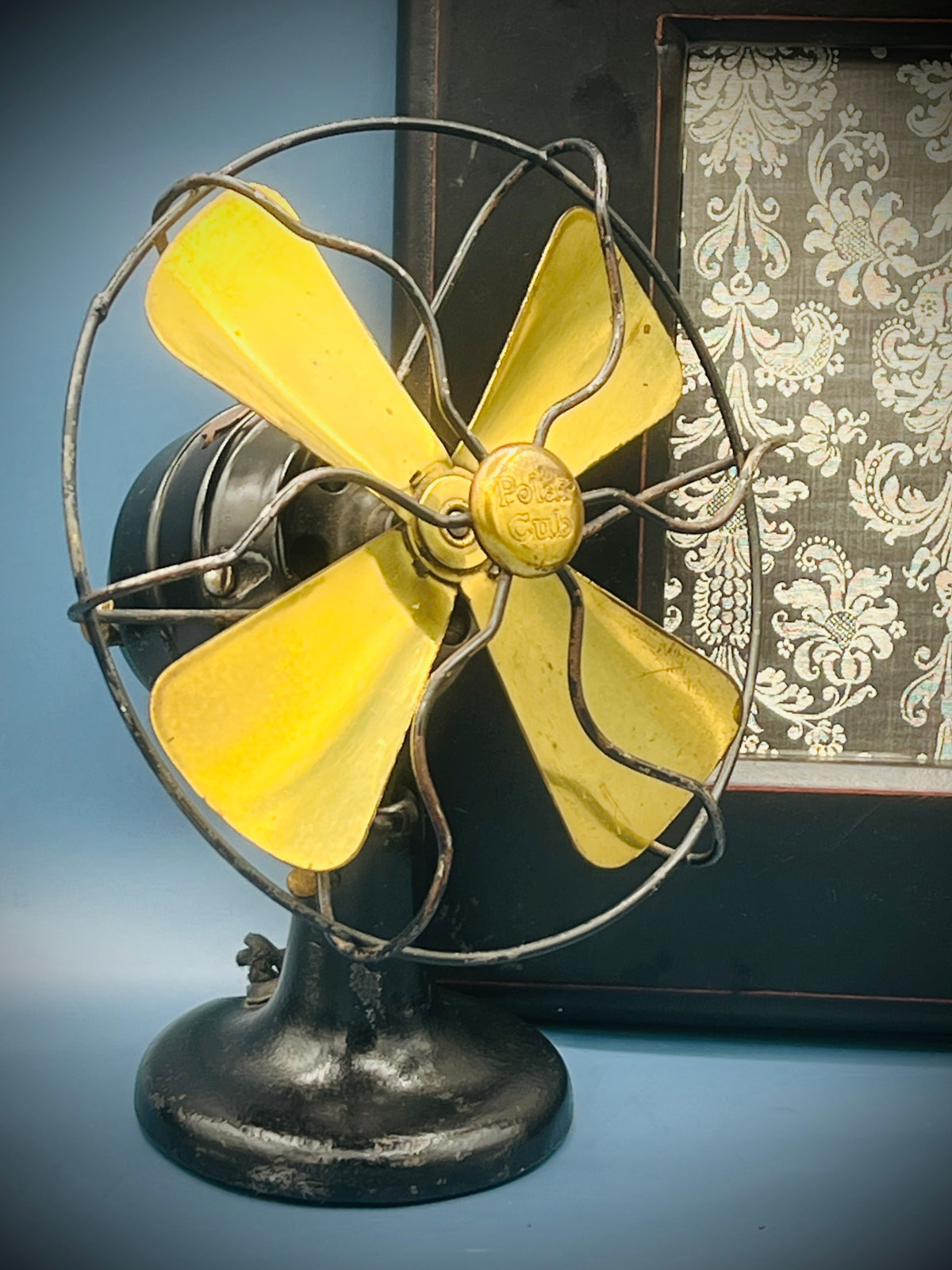 A vintage A.C. Gilbert Polar Cub electric fan featuring four brass blades, a wire cage guard, and a black painted metal base set against a patterned background. - Photo 3
