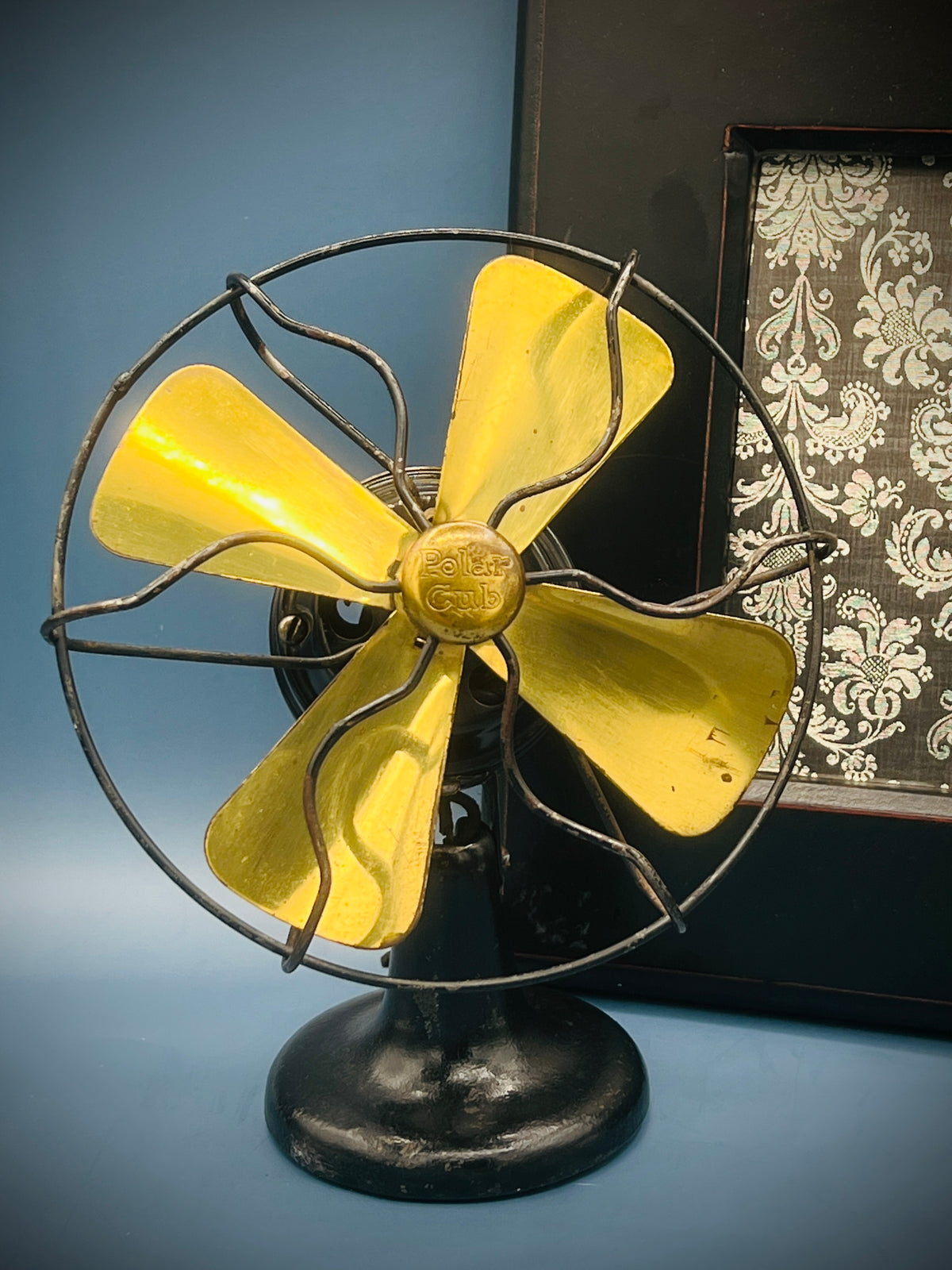 A vintage A.C. Gilbert Polar Cub electric fan featuring four brass blades, a wire cage guard, and a black painted metal base set against a patterned background. - Photo 4