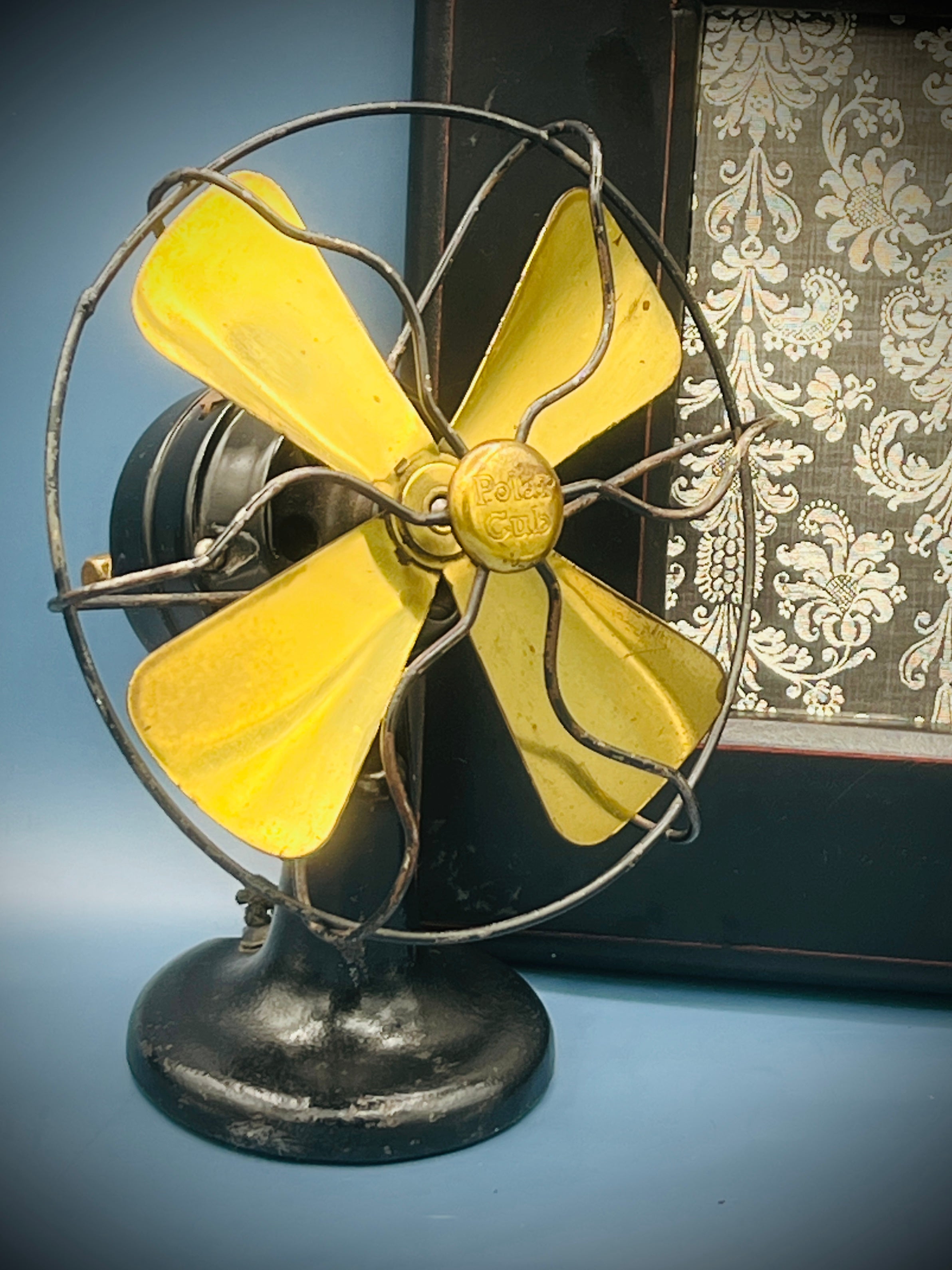 A vintage A.C. Gilbert Polar Cub electric fan featuring four brass blades, a wire cage guard, and a black painted metal base set against a patterned background. - Photo 5