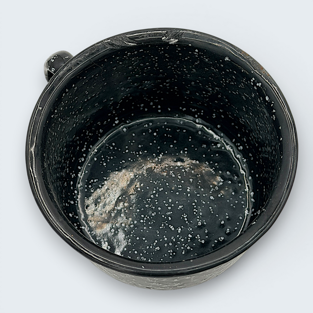 A vintage black enamel cup with white speckles and a single handle, showing a rustic interior with character-building wear at the bottom. - Photo 6