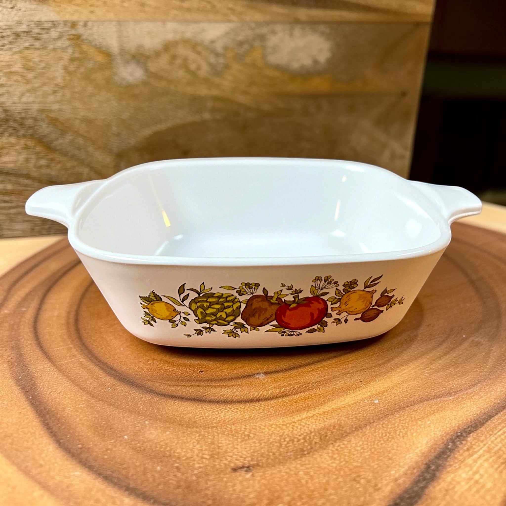 A square-shaped white Corning Ware petite casserole dish with lug handles, resting on a rustic wood slice background. - Photo 1