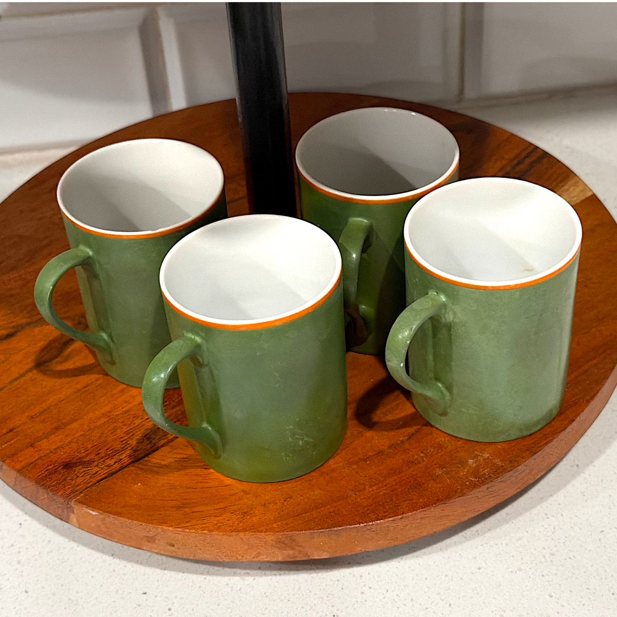 A mid-century modern cylindrical green porcelain mug featuring a bold orange stripe along the upper rim, a white interior, and an attached side handle, standing upright on a round wooden surface. - Photo 1