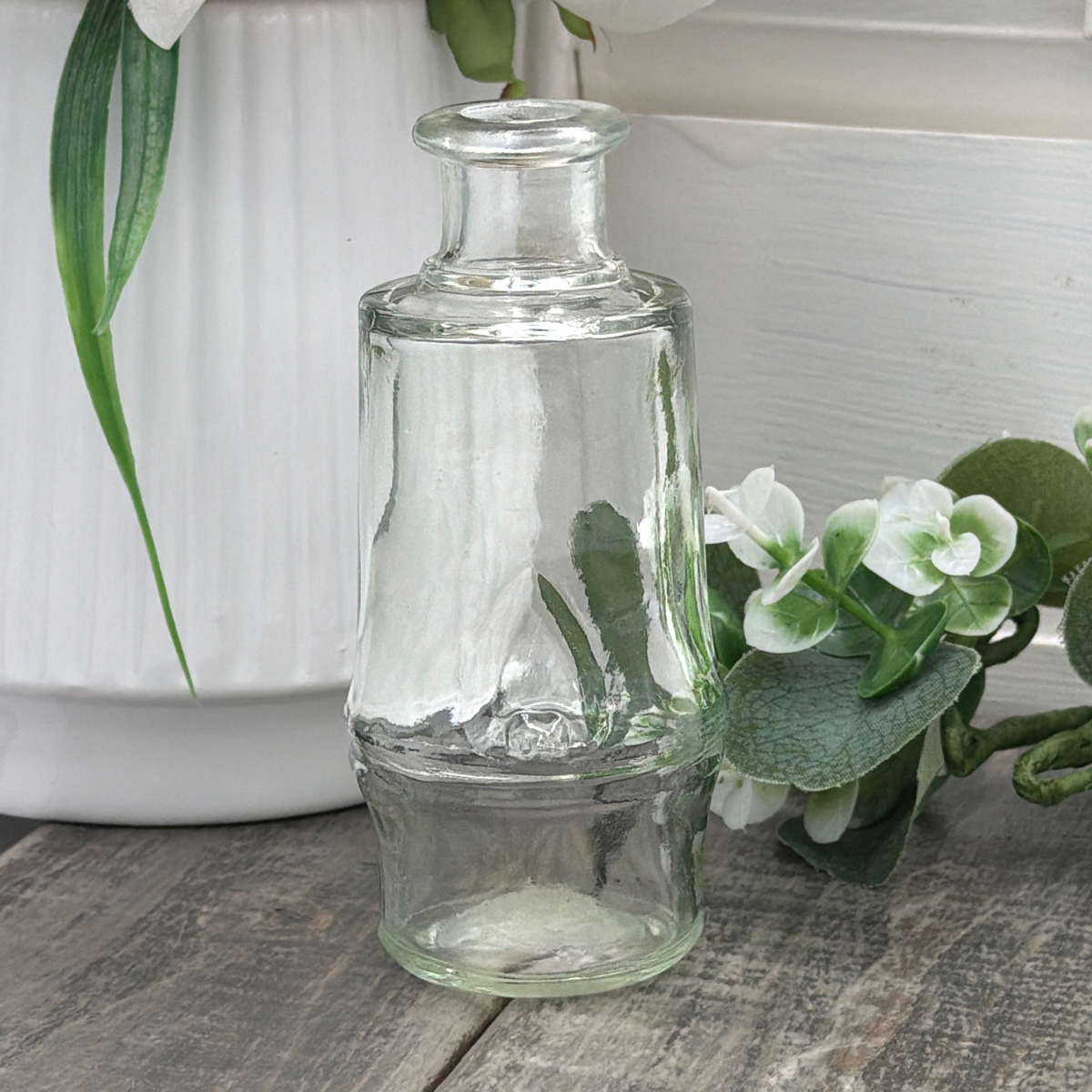 Vintage clear glass cylindrical bottle with a flared rounded rim, raised central horizontal ridge, and subtly tapered base, displayed upright beside green leaves and a white vase on a weathered wood surface. - Photo 1