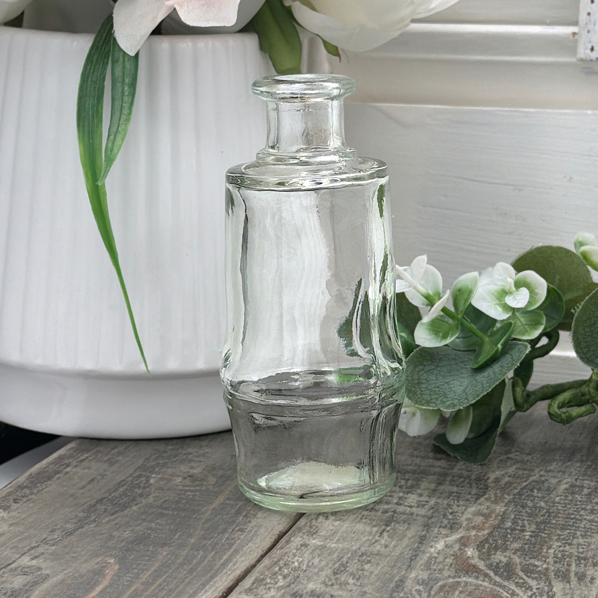 Vintage clear glass cylindrical bottle with a flared rounded rim, raised central horizontal ridge, and subtly tapered base, displayed upright beside green leaves and a white vase on a weathered wood surface. - Photo 2