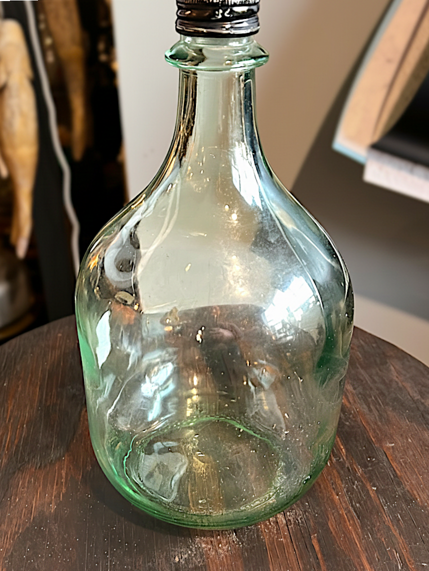 Vintage pale green glass one-gallon jug with bulbous body, fluted shoulder, and black metal screw cap resting on a dark wood surface. - Photo 1