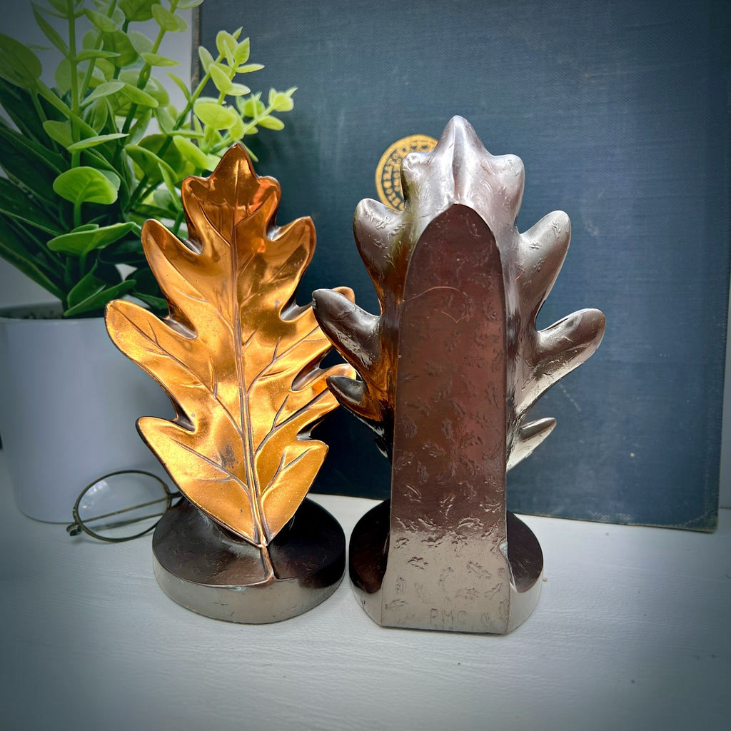 A pair of vintage PMC hand-cast metal oak leaf bookends with a rich bronze finish, designed with stylized lobed leaves and delicate embossed cascading leaf patterns on their curved backs, supporting a dark book on a white surface. - Photo 2