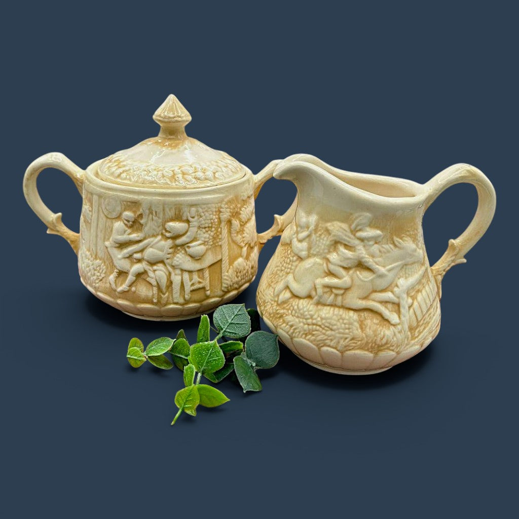 A cream-colored antique ceramic creamer pitcher featuring detailed relief-molded scenes of tavern figures under a smooth glazed finish. - Photo 1