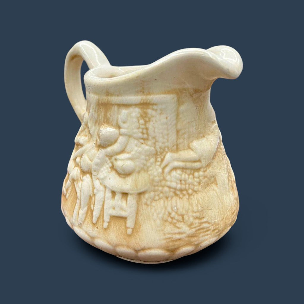 A cream-colored antique ceramic creamer pitcher featuring detailed relief-molded scenes of tavern figures under a smooth glazed finish. - Photo 6